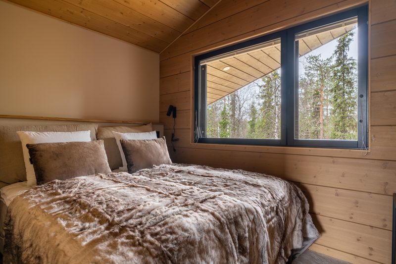 A bedroom with a double bed made up in linen and fur, beside a long low window onto the trees.