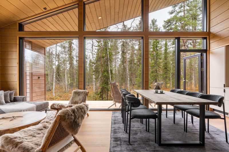 The living room with the glass door slid open onto the deck, the dining table beside it and forest beyond.