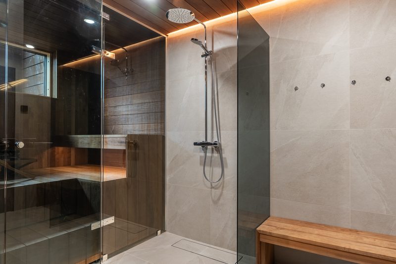 The washroom with a rain shower behind glass, stone-effect tiles, and the sauna visible through the glass wall.