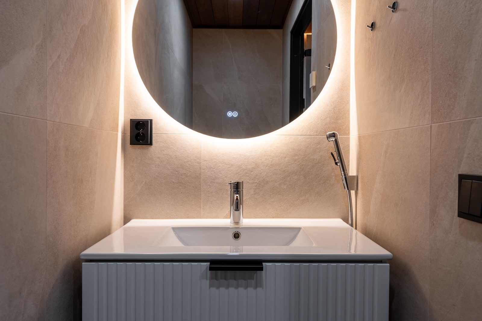 A separate WC with a round backlit mirror, pale ribbed vanity and stone-effect tiles.
